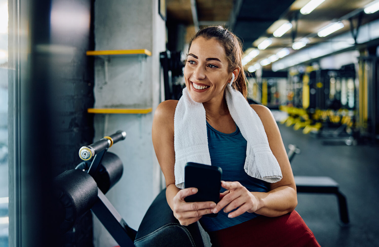 Happy sportswoman using smart phone while listening music over earbuds in health club and looking away. Copy space.